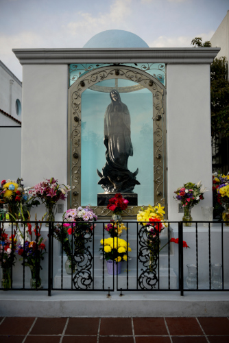 A picture of Mother Mary statue framed on a wall with flowers placed infront of it 
